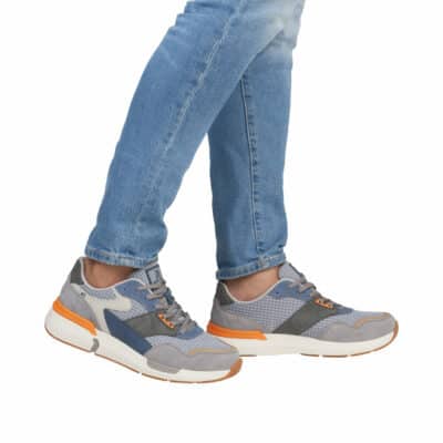 En person går i Rieker Sneakers Herre og viser gråt atletisk design med orange og hvide detaljer, parret med lyseblå jeans mod en almindelig hvid baggrund. Fokus er på deres ben og fødder.