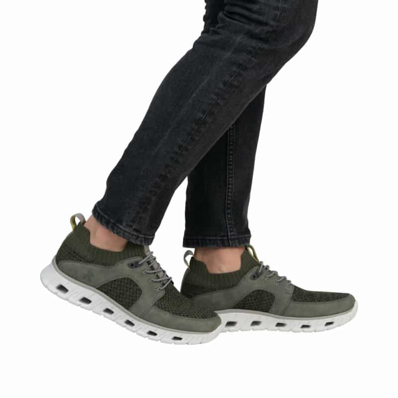 En person går på en hvid baggrund i Rieker Sneakers Herre, med olivengrønne overdele med hvide såler, parret med mørke jeans.